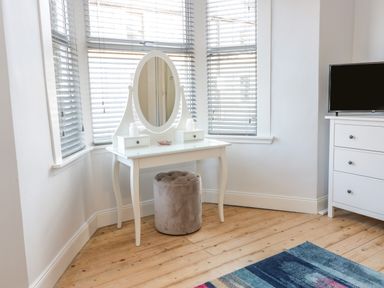 A room with a white vanity table with an oval mirror a gray ottoman a white dresser with a television and wooden floors at The Lazy Seal in Spittal