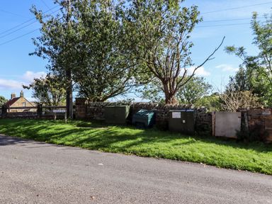 A street view with grass and trees alongside three green electrical boxes and a stone wall at Michaelmass Cottage in Castleton