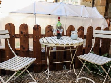 A small outdoor seating area with two white metal chairs and a round table with a bottle and two glasses on the table at Michaelmass Cottage in Castleton