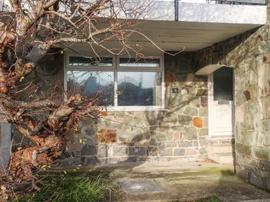 The exterior of a stone house with a white door a window and a tree in front at 5 Ger Y Nant in Llanbedrog