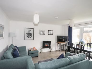 A living room with blue sofas a television a dining table and windows at 5 Ger Y Nant in Llanbedrog