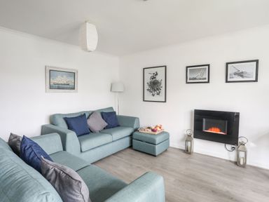 A living room with blue sofas cushions an ottoman wall art and a wall-mounted electric fireplace at 5 Ger Y Nant in Llanbedrog