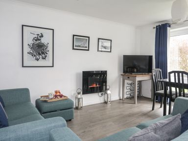 A living room with blue couches a wall-mounted fireplace a television on a corner table and framed pictures on the wall at 5 Ger Y Nant in Llanbedrog