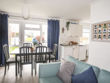 A dining area with a table and six chairs next to a kitchen with white cabinets and a light blue sofa in Llanbedrog