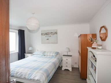 A bedroom with a double bed between two bedside tables with lamps a wardrobe a white bookshelf and a window with dark curtains at 5 Ger Y Nant in Llanbedrog