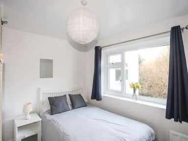 A bedroom with a double bed two pillows a bedside table with a lamp and a window with dark curtains and flowers on the sill at 5 Ger Y Nant in Llanbedrog
