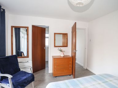 A bedroom with a wooden chest of drawers with a mirror above it two open wooden doors and a chair with a dark cover at 5 Ger Y Nant in Llanbedrog