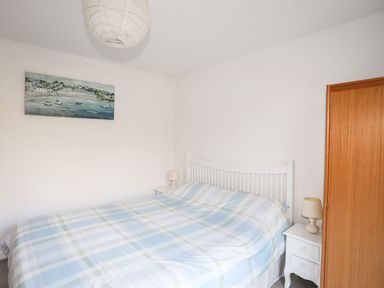 A bedroom with a bed covered in a blue and green checkered blanket two white bedside tables with lamps a wooden door and a painting above the bed at 5 Ger Y Nant in Llanbedrog