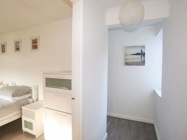A bedroom with a bed next to a white nightstand and drawers and a hallway with a hanging light and wall art at 5 Ger Y Nant in Llanbedrog