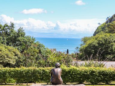 A bronze statue of a seated person looking towards the sea with green bushes and hills in the background at 5 Ger Y Nant in Llanbedrog