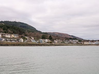 A coastal town with houses along a rocky shoreline and forested hills in the background at Rocklea Seaside Cottage in Newcastle County Down