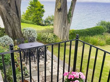 A garden terrace with a metal table and chairs near large trees overlooking the sea at Rocklea Seaside Cottage in Newcastle County Down