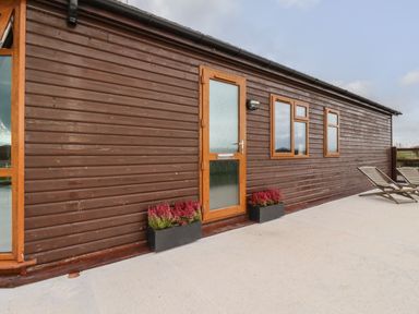 The exterior side of a wooden lodge with a door, two windows, flower boxes, and two lounge chairs on a concrete patio