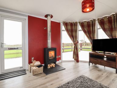 A room with a wood burning stove next to a door and windows with plaid curtains and a television on a wooden stand at Sycamore Lodge in Porkellis near Helston