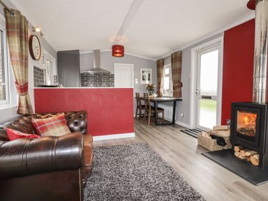 A living area with a brown leather sofa and plaid pillows a wood burning stove with a basket of firewood and a dining table with chairs at Sycamore Lodge Porkellis near Helston