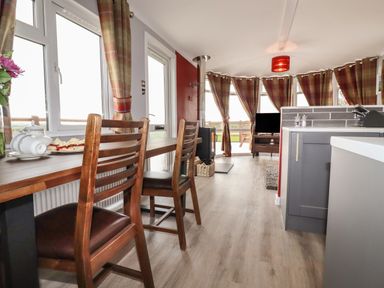 A dining area with wooden chairs and table next to windows and a kitchen with grey cabinets at Sycamore Lodge in Porkellis near Helston