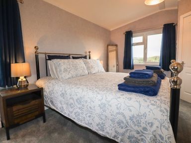 A bedroom with a bed with patterned bedding a side table with a lamp curtains and towels on the bed at Sycamore Lodge in Porkellis near Helston