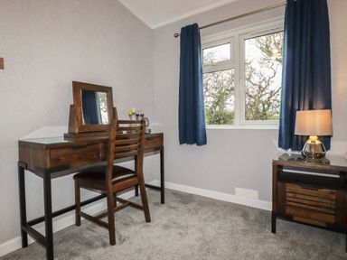 A room with a wooden desk and chair next to a window with blue curtains and a wooden side table with a lamp at Sycamore Lodge in Porkellis near Helston