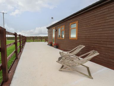 An outdoor patio with wooden lounge chairs beside a wooden building with windows and door at Sycamore Lodge Porkellis near Helston