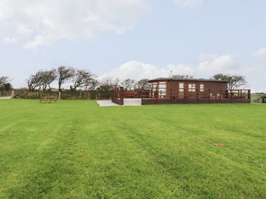 A wooden lodge with a fenced deck on a grassy field with trees at Sycamore Lodge in Porkellis near Helston