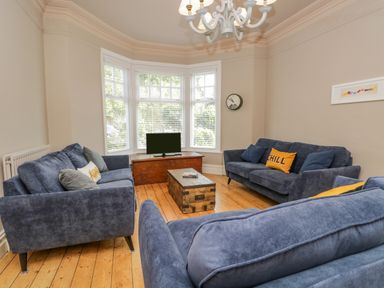 A living room with three blue sofas a wooden floor a wooden chest used as a coffee table a TV on a low wooden cabinet and large bay windows at Hollywood in Windermere