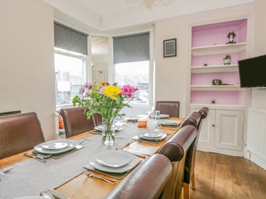 A dining room with a wooden table set for six with plates and cutlery and a vase of flowers by windows at Hollywood in Windermere