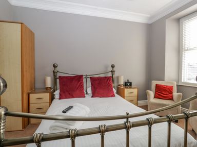 A bedroom with a metal bed frame white bedding red pillows two wooden nightstands lamps a beige armchair with a red cushion near a window with blinds and a wooden wardrobe at Hollywood in Windermere