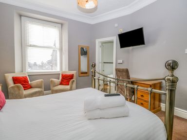 A bedroom with a metal bed frame two beige armchairs with red cushions a wooden desk and chair and a wall-mounted television at Hollywood in Windermere