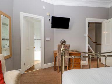A bedroom with a metal bed frame a wooden desk with a chair a wall-mounted TV an armchair a mirror and two open doors one leading to a bathroom and the other to a hallway at Hollywood in Windermere