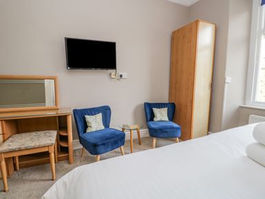 A bedroom with a wall mounted TV two blue chairs a small glass table a wooden wardrobe and a wooden vanity with a stool at Hollywood in Windermere
