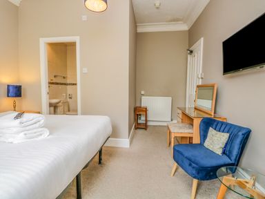 A bedroom with a bed and towels a blue chair a dressing table with a mirror and a wall-mounted tv at Hollywood in Windermere