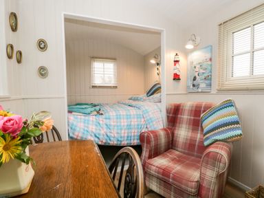 A room with a wooden table and chairs a red plaid armchair a bed with blue checked bedding and windows with blinds at Seashore Shepherds Hut @ Moat Farm in Aldingham near Ulverston