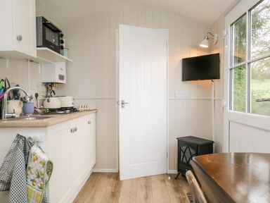 A small kitchen area with cabinets a microwave kettle and toaster a door a wall mounted television a small stove and a table near a window at Seashore Shepherds Hut @ Moat Farm in Aldingham near Ulverston