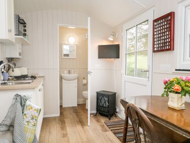 A kitchen and dining area with a wooden table flowers on the table a small stove and a door leading to a bathroom at Seashore Shepherds Hut @ Moat Farm Aldingham near Ulverston