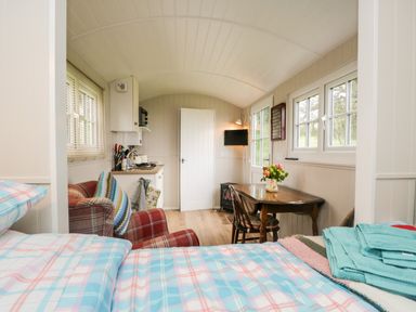 An interior view of a small room with a bed in the foreground a plaid armchair a wooden table with chairs a small kitchen area and windows at Seashore Shepherds Hut @ Moat Farm Aldingham near Ulverston