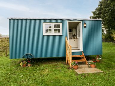 A blue corrugated metal shepherds hut with wooden steps and potted flowers on grass at Seashore Shepherds Hut @ Moat Farm in Aldingham near Ulverston