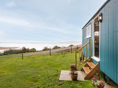 A blue shepherds hut with wooden steps and potted plants on grass near a gravel path at Seashore Shepherds Hut @ Moat Farm in Aldingham near Ulverston