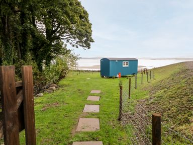 A blue shepherds hut on grass near a fenced area with trees and a view of water at Seashore Shepherds Hut @ Moat Farm in Aldingham near Ulverston