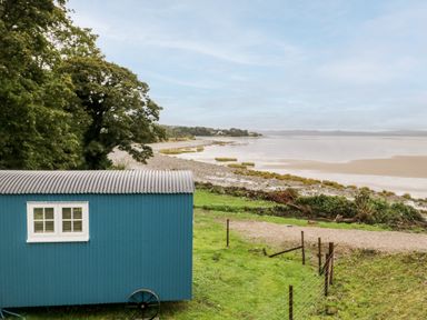 A blue shepherds hut near a grassy area by a rocky coastline with trees in the background at Seashore Shepherds Hut @ Moat Farm in Aldingham near Ulverston