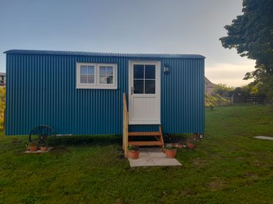 A blue shepherds hut with a white door and window on grass with flower pots in Aldingham near Ulverston