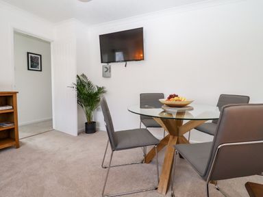 A dining area with a glass table and gray chairs a wall-mounted tv a plant and a wooden shelf at 3 Granbrook Lane in Mickleton Gloucestershire