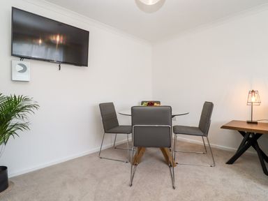 A dining area with a glass table and four grey chairs a wall-mounted television a small plant and a side table with a lamp at 3 Granbrook Lane in Mickleton Gloucestershire