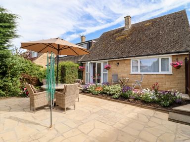 A stone patio with an outdoor table and chairs under a large umbrella and flowerbeds by the house at 3 Granbrook Lane in Mickleton Gloucestershire