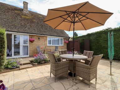 An outdoor patio area with a round table, four wicker chairs, a large umbrella, flowers, and a house with windows at 3 Granbrook Lane Mickleton Gloucestershire