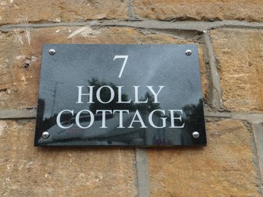 A black house number sign with white text reading 7 Holly Cottage on a stone wall at Holly Cottage in West Coker