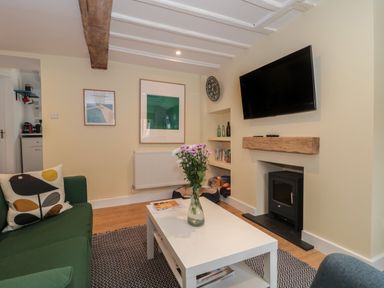 A living room with a green sofa a white coffee table with flowers a wall-mounted television and a small stove at Holly Cottage in West Coker