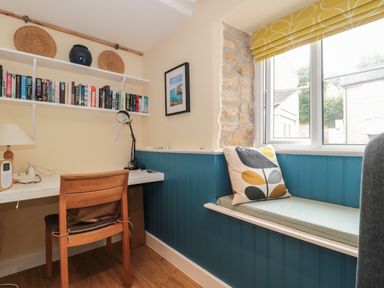 A small study area with a wooden chair a desk with a lamp and books on shelves next to a window seat with a cushion at Holly Cottage in West Coker