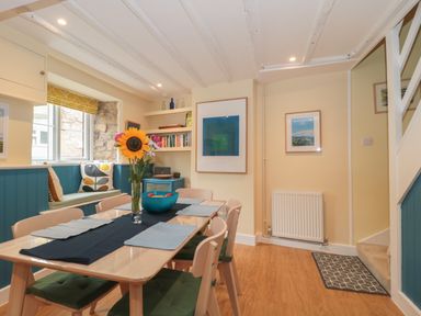 A dining area with a wooden table and chairs, a vase with flowers, blue cushions on window seating, and framed pictures on walls at Holly Cottage in West Coker
