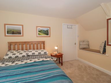 A bedroom with a wooden bed a bedside table with a lamp a window seat with cushions and framed pictures on the wall at Holly Cottage in West Coker