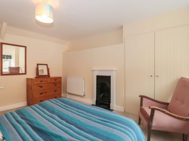 A bedroom with a blue striped bedspread a wooden chest of drawers with mirrors a fireplace a radiator a pink armchair and built in cupboards at Holly Cottage in West Coker